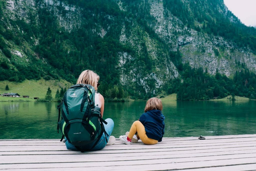 Mère et fille randonneuses de dos avec sac à dos assis sur un ponton en bois admirant le lac Königssee et les montagnes de Bavière en famille