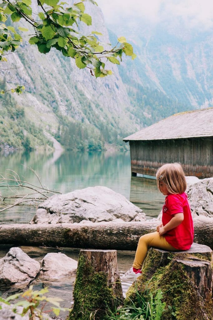 Jeune enfant assise sur une souche de bois admirant les montagnes et le reflet de la forêt dans le lac Obersee en Allemagne face a la cabane de pêcheur