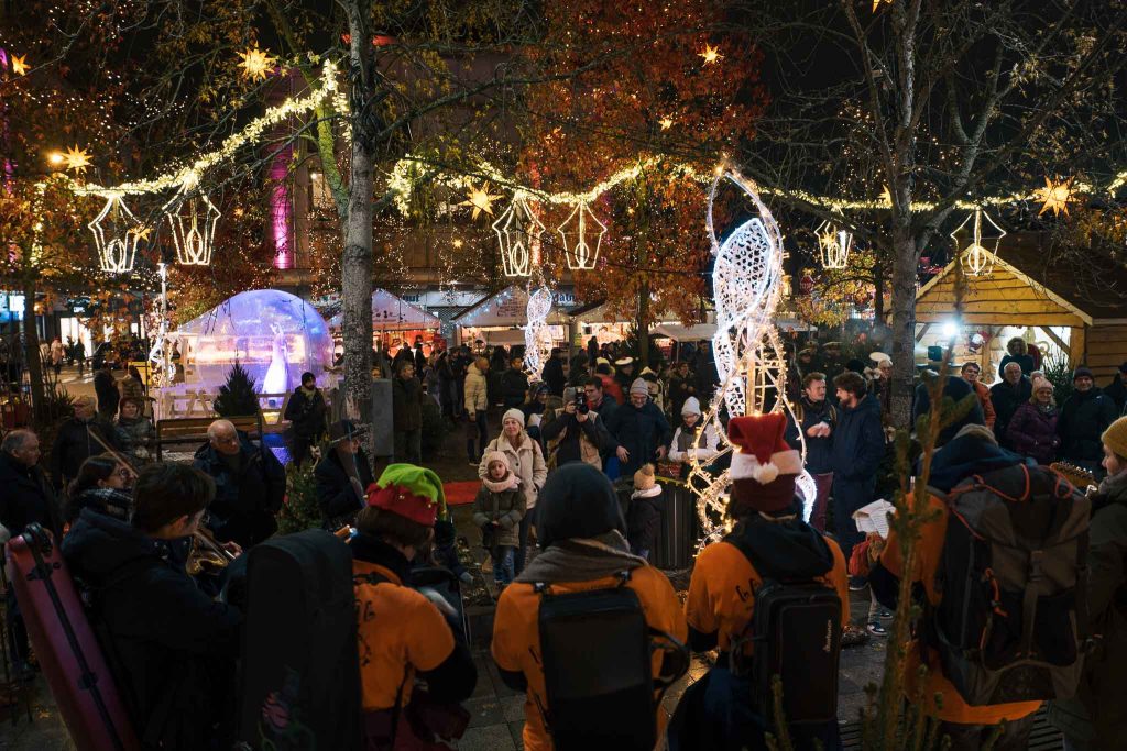 Vue d'ensemble de l'ambiance féerique du marché de Noël et des illuminations à Épinal, avec des musiciens en tenue de fête au premier plan.
