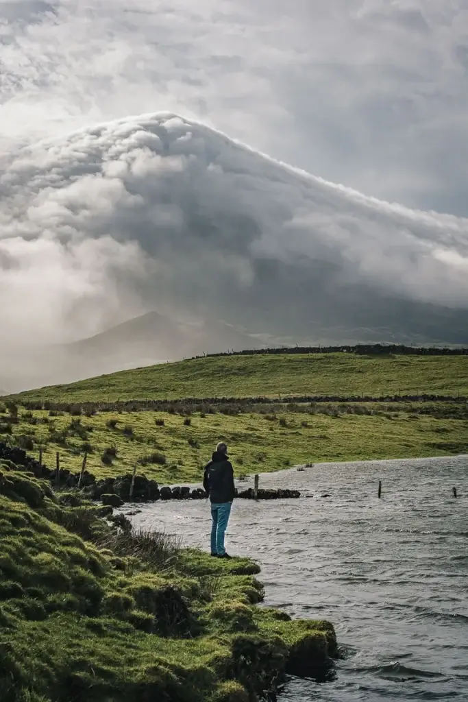 Homme debout au bord d'un lac face au volcan Pico partiellement dégagé, île de Pico aux Açores