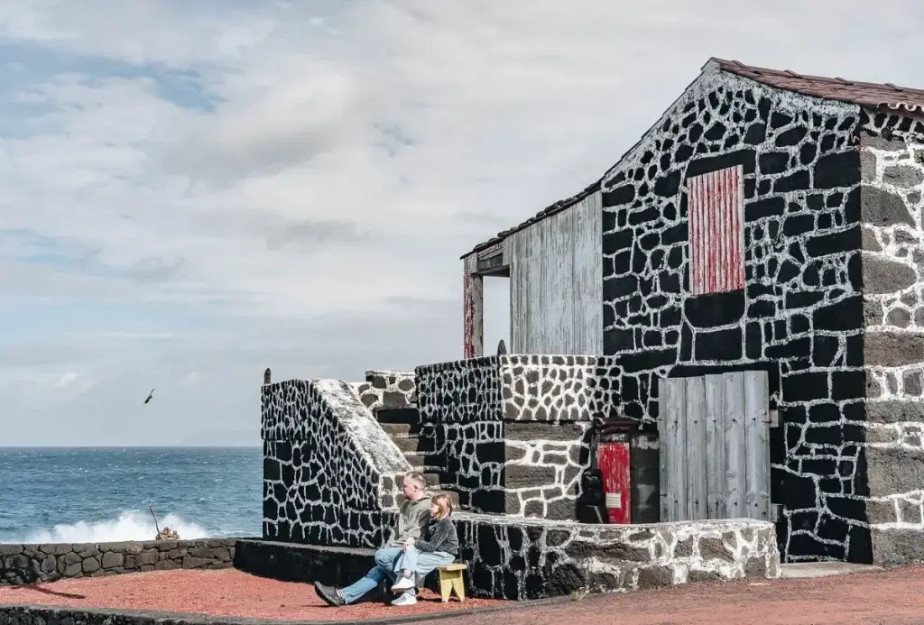 Père et fille assis devant une maison en pierre de lave noire et blanche au bord de la mer à Lajido, île de Pico aux Açores